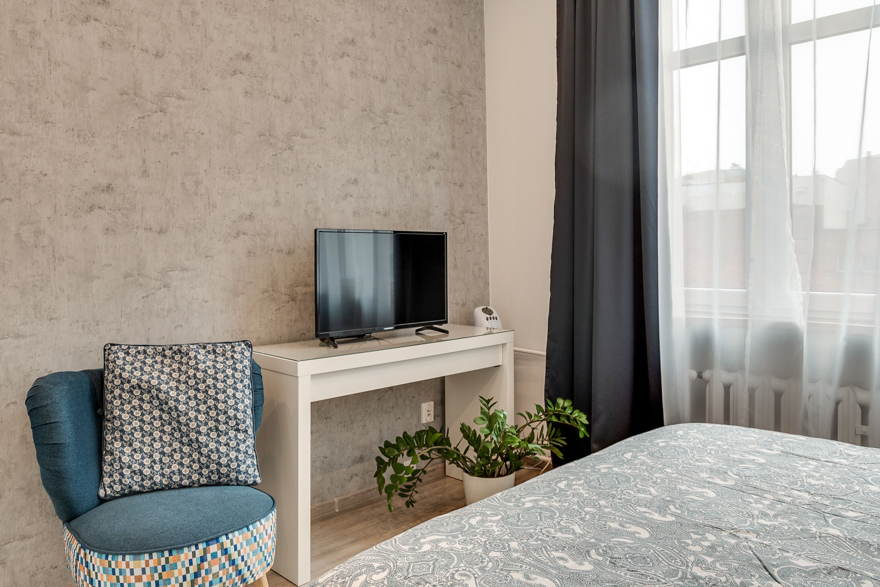 Habitació d'hotel moderna amb un televisor de pantalla plana sobre una taula de nit blanca, un sofà blau amb coixins estampats, un llit amb edredó estampat i vista a edificis a través de cortines primes.