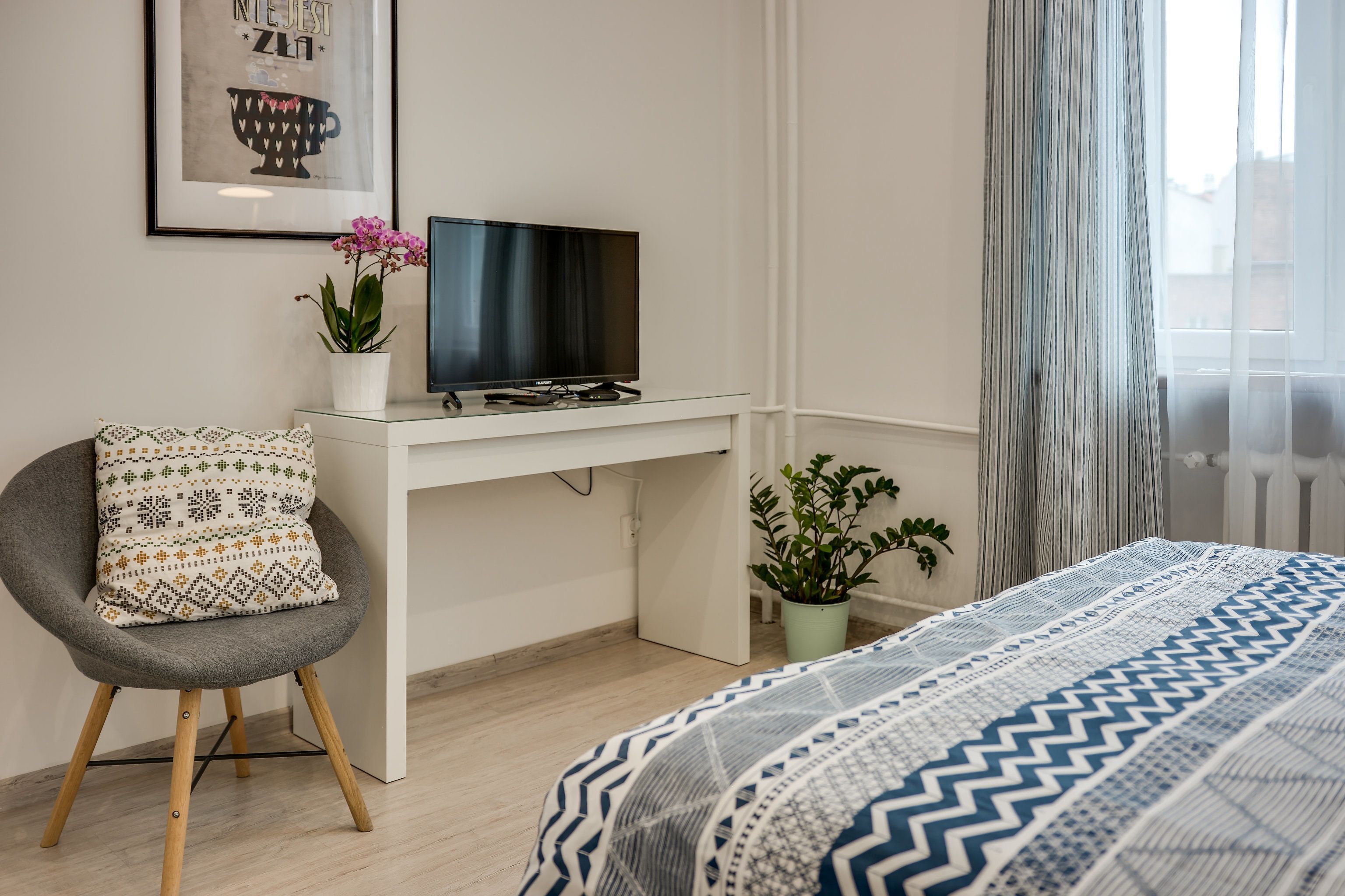 Quarto de hotel moderno com uma escrivaninha branca, televisão, poltrona cinza e cama com colcha estampada.