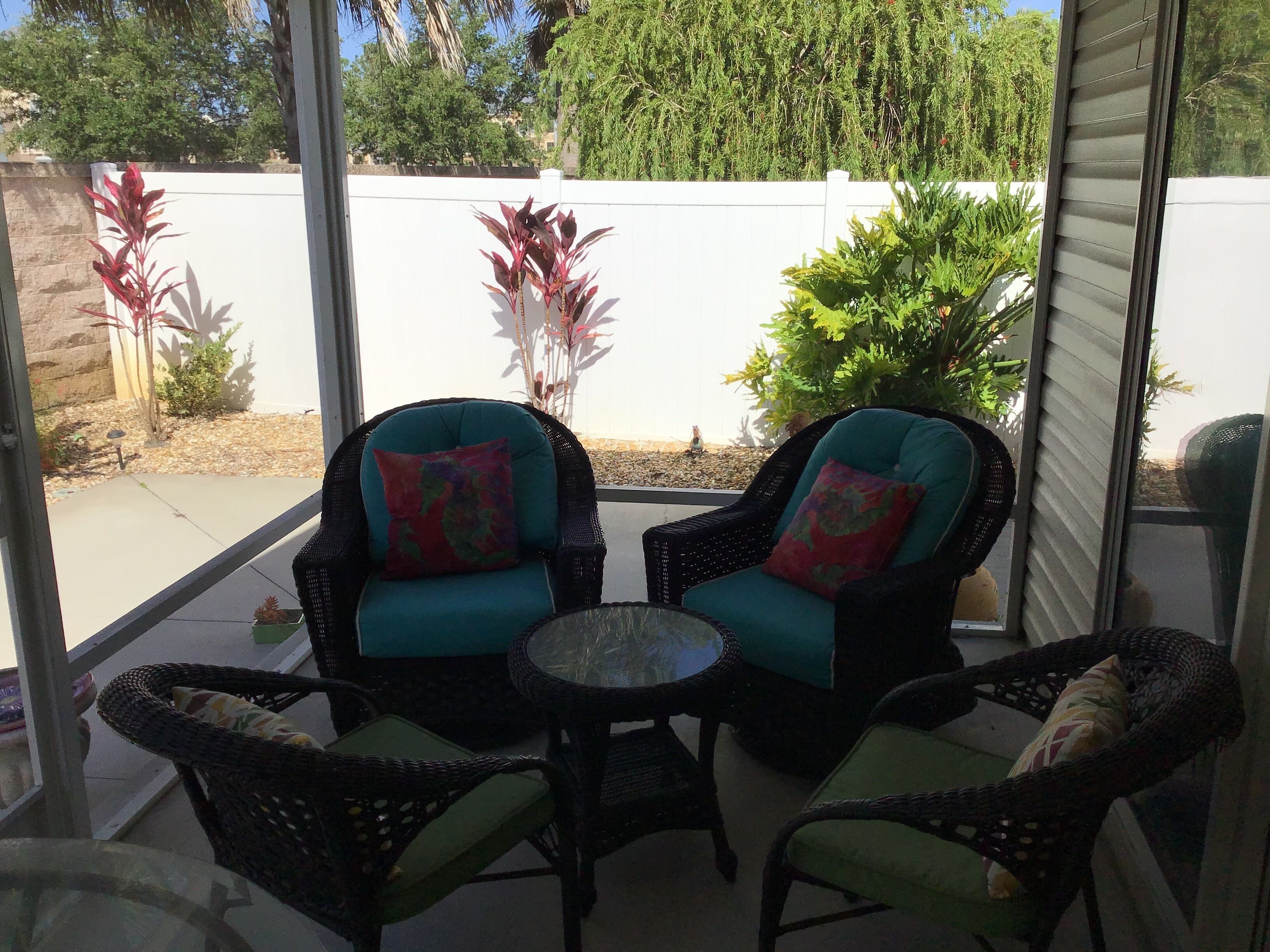 A covered patio area with wicker furniture and a small table, surrounded by potted plants and a white fence.