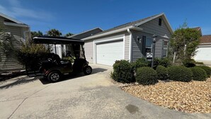 A residential garage with a white garage door, a black golf cart parked in the driveway, and a well-maintained lawn with trimmed bushes. - 3-Bed Dog-Friendly Rental: Near Brownwood Paddock & Outdoor Oasis (The Villages)