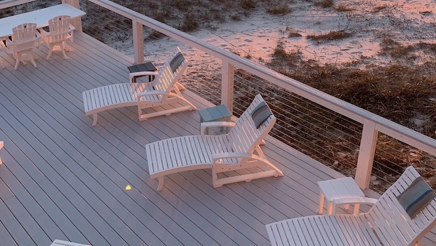 Un terrazzo in legno con poltrone da relax bianche e un tavolo, con vista su una spiaggia al tramonto.