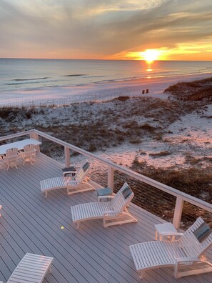 Eine Holzterrasse mit weißen Liegestühlen und einem Tisch, mit Blick auf einen Strand bei Sonnenuntergang.