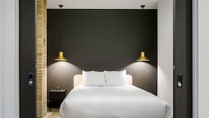 A modern bedroom with a large bed, two wall-mounted lamps, a small bedside table, and a brick accent wall. - Les Lofts de la Gare - By Les Lofts Vieux-Quebec (Québec City)