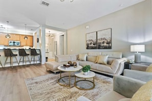A modern living room with a sofa, coffee table, and chairs. There is a kitchen in the background.