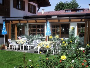 An outdoor dining area with tables and chairs, surrounded by a garden with flowers and plants. - Hotel Feldmeier (Oberammergau)