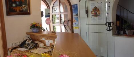 A reception area with a wooden desk, a telephone, and a vase of flowers. There is a framed painting on the wall and a door with a coat of arms.