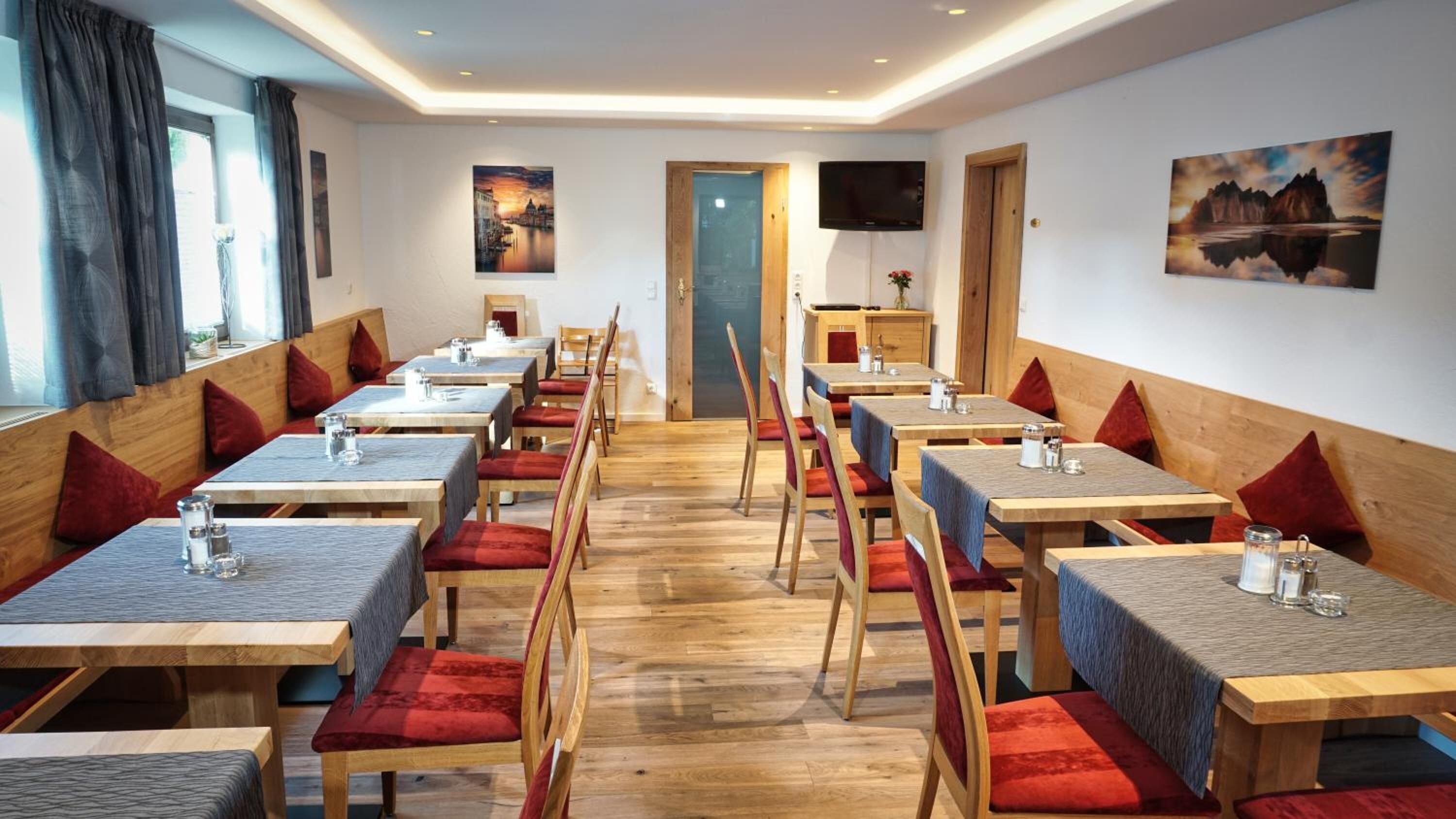 A dining area with wooden tables and red cushioned chairs, a TV mounted on the wall, and artwork on the walls.