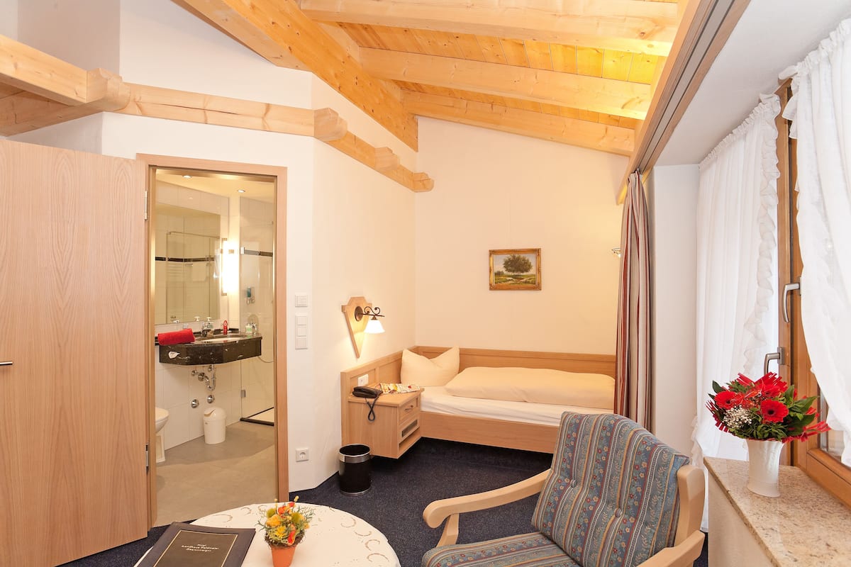 Een kleine, knusse kamer met een bed, een stoel, een nachtkastje en een spiegel, plus een raam met gordijnen.