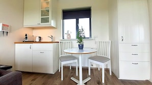 Una cocina compacta con armarios blancos, una pequeña mesa de comedor con dos sillas, una planta en maceta y una ventana con vistas.