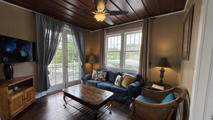 A cozy living room with a wooden ceiling, a sofa with cushions, a wicker chair, a coffee table, and a television on the wall. - Park Place Inn and Cottages (Sanford)