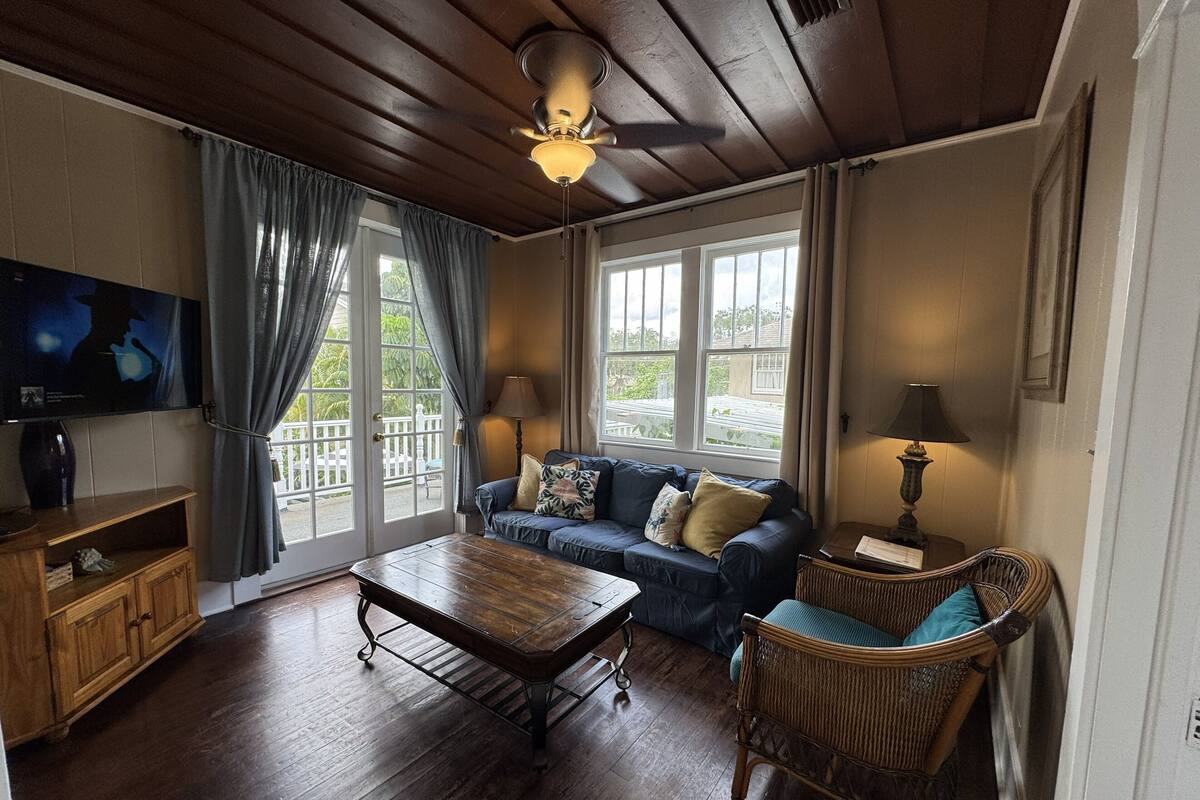 A cozy living room with a wooden ceiling, a sofa with cushions, a wicker chair, a coffee table, and a television on the wall.