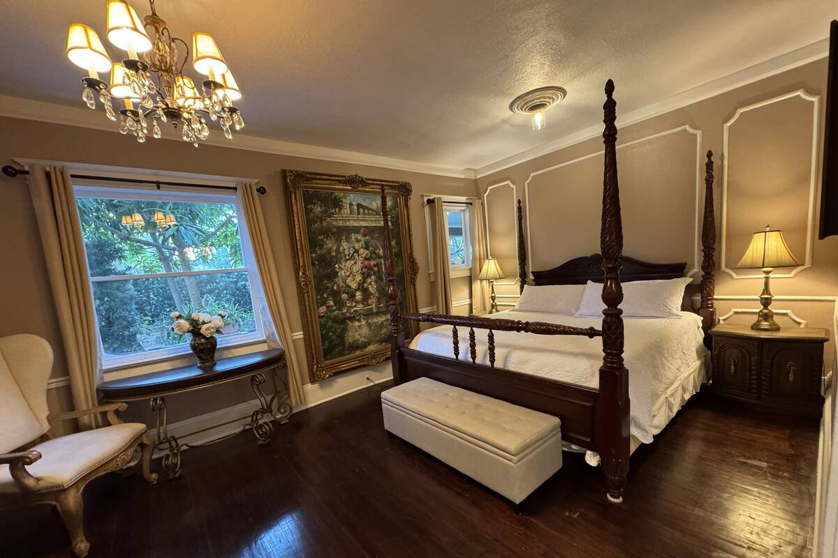 A four-poster bed with a canopy, a sitting bench, a chandelier, and a large window with curtains.