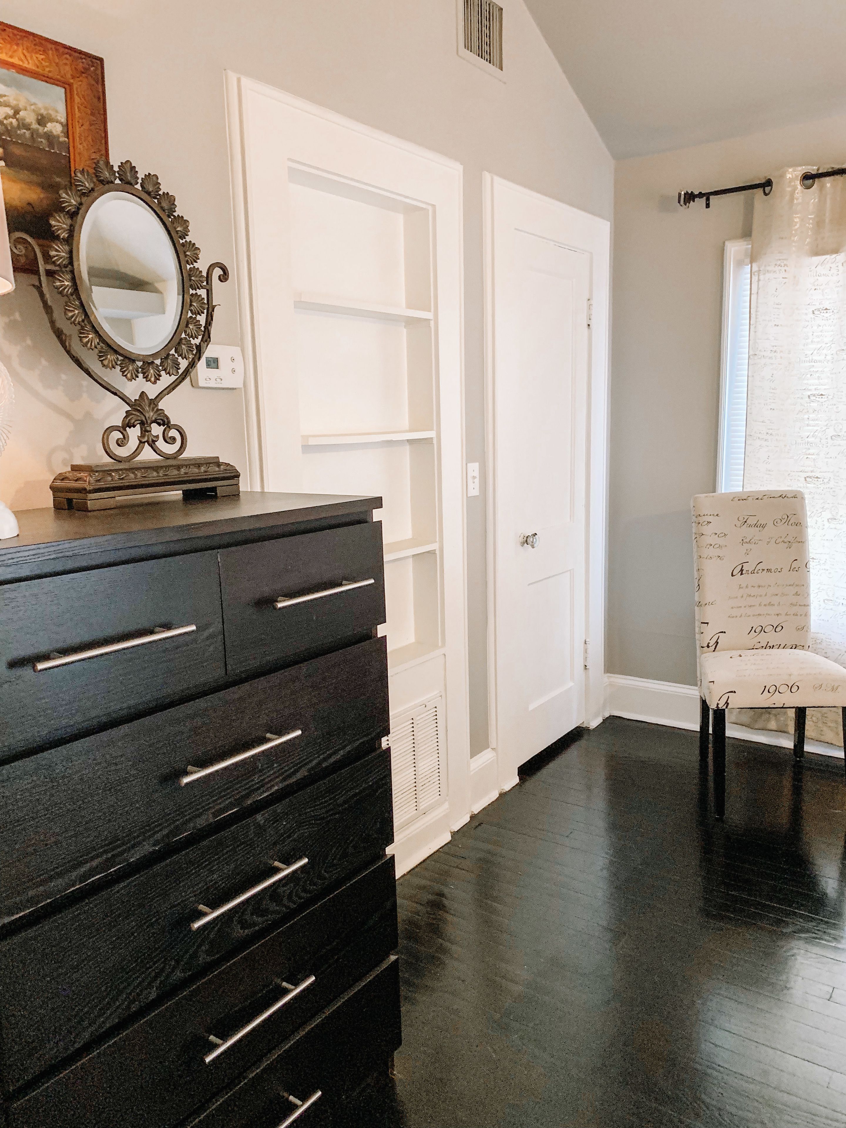 A room with a black dresser, white doors, a mirror with a decorative frame, and a patterned chair.