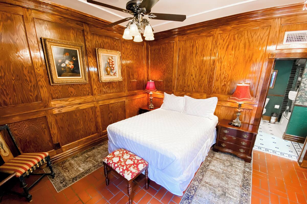 A hotel room with a large bed, wooden paneling, a ceiling fan, and a bathroom visible through an open door.