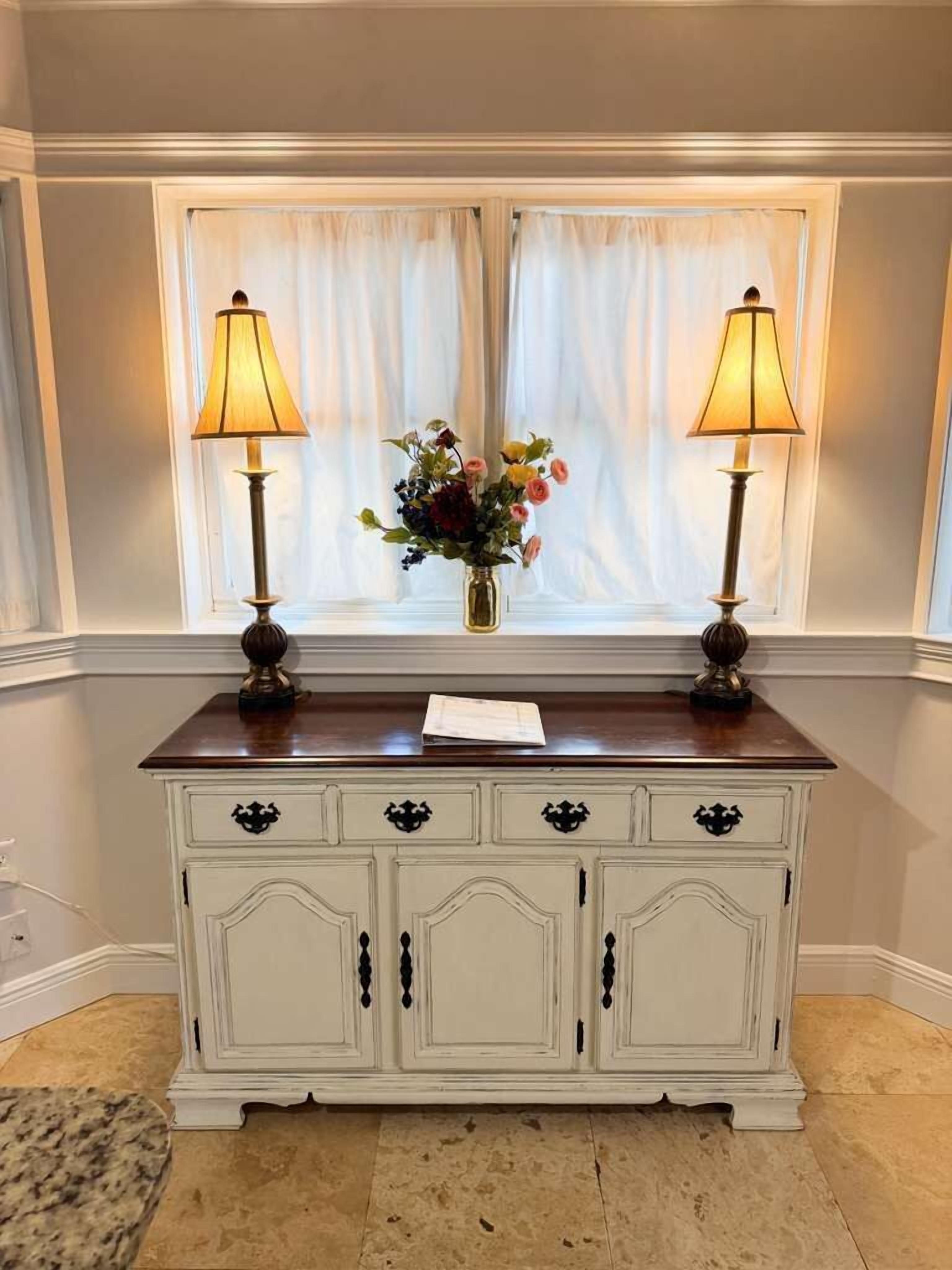 A white dresser with two lamps, a vase with flowers, and a window with curtains.