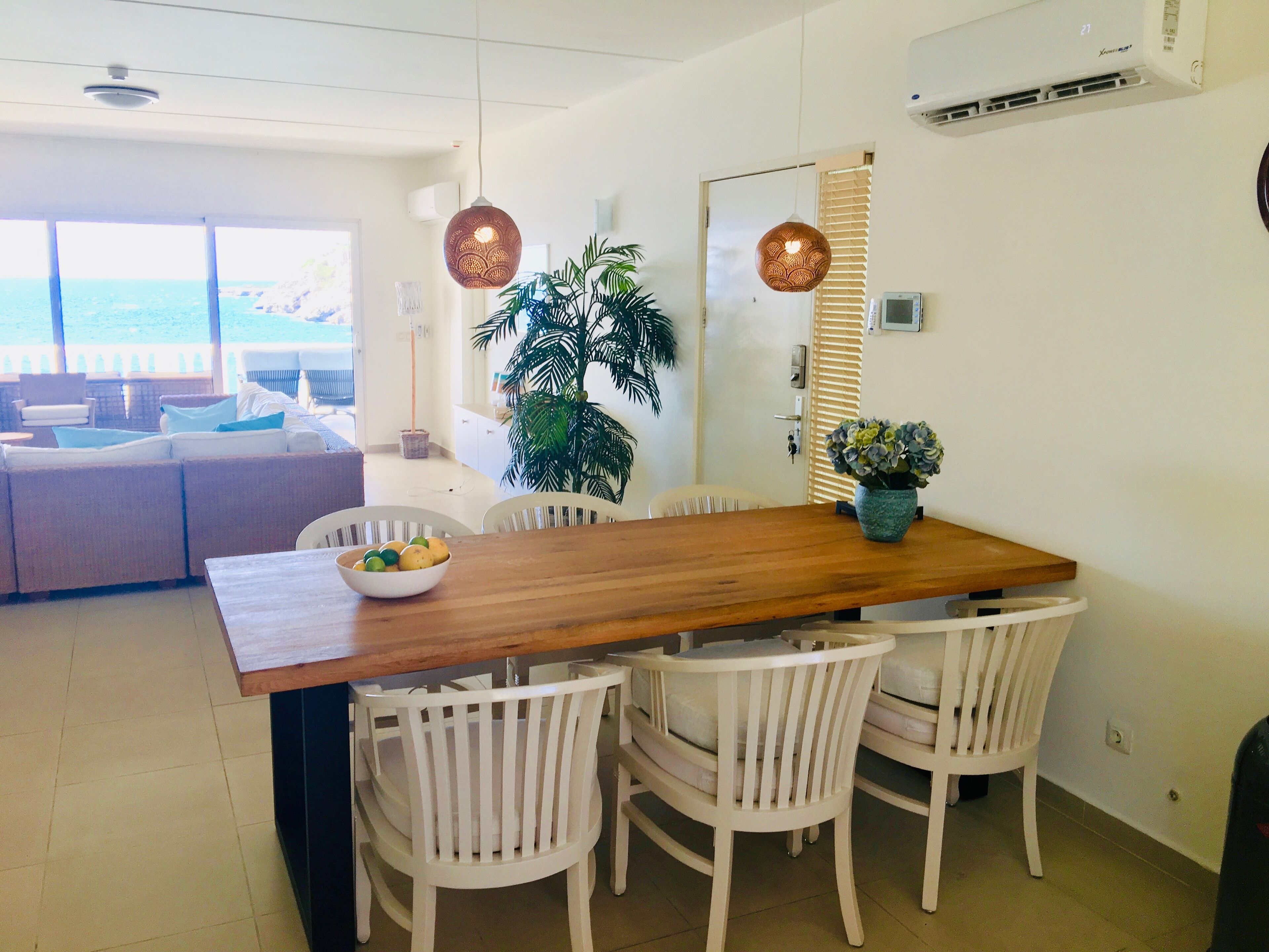 A dining area with a wooden table, white chairs, a bowl of fruit, and a potted plant.