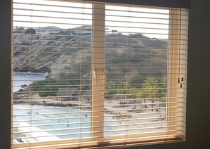 A window with horizontal blinds, a view of a beach with people and boats, and a rocky hillside in the background.