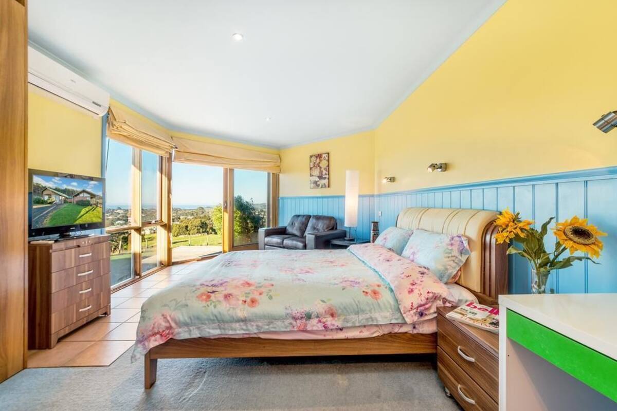 A bedroom with a large bed, a TV, a desk, and a view of the outdoors.