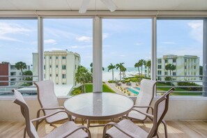 Una habitación con una mesa redonda y cuatro sillas, ventanas amplias que dan a una playa y edificios, y una zona de piscina visible a través de las ventanas.