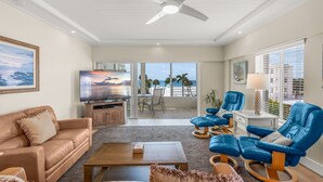 A spacious living room with a ceiling fan, a flat-screen TV, a sofa, a coffee table, and a balcony with a view of the ocean. - Island House Beach Resort 13S (Siesta Key)