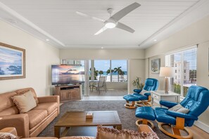 A spacious living room with a ceiling fan, a flat-screen TV, a sofa, a coffee table, and a balcony with a view of the ocean.