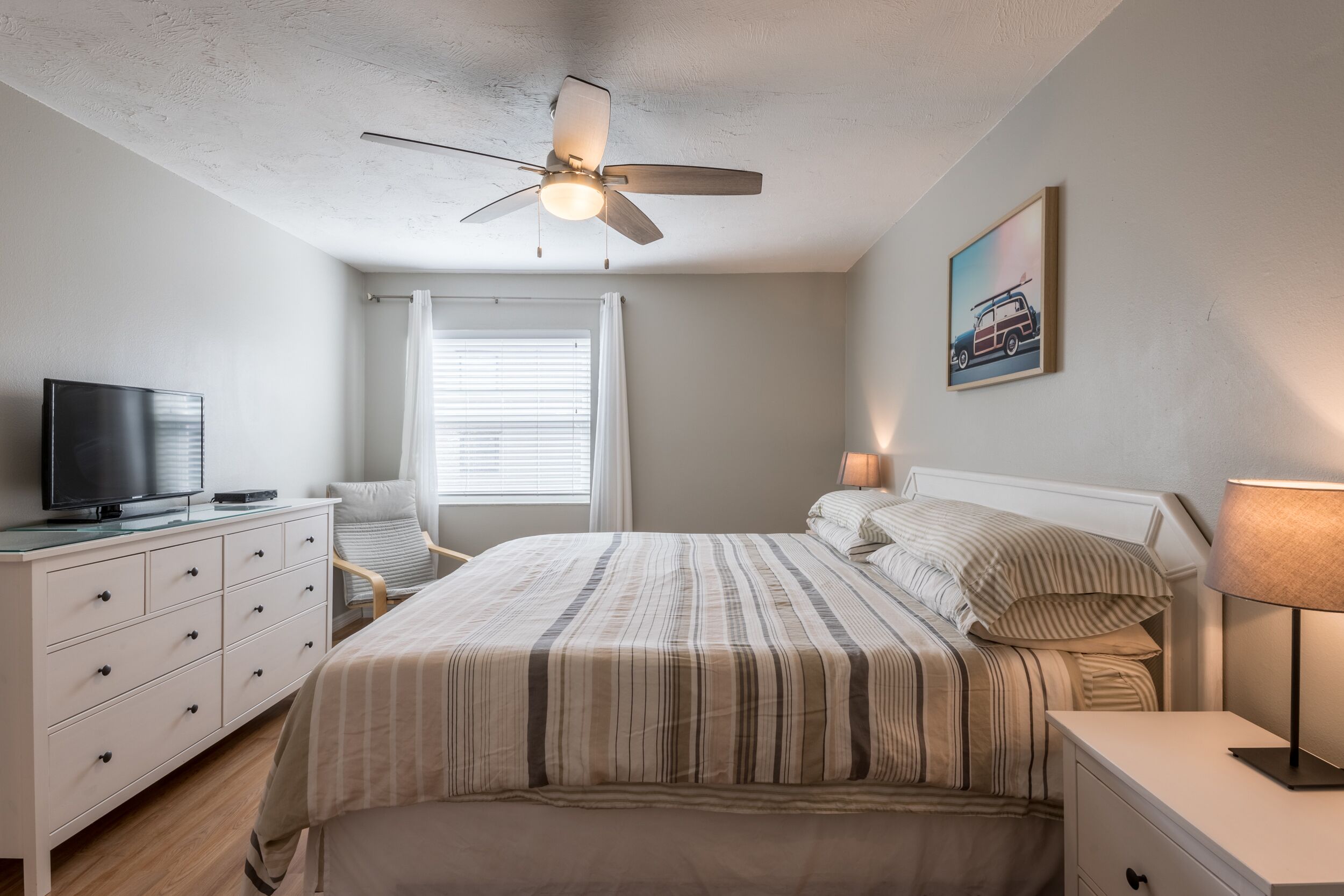 A bedroom with a bed, a TV on a dresser, a ceiling fan, a window with blinds, and a wall-mounted lamp.