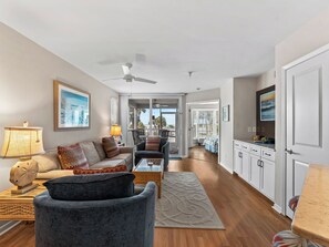 A spacious living area with a sofa, armchairs, a coffee table, and a TV. There is a ceiling fan, a window with a view, and a door leading to another room.