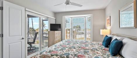 A bedroom with a bed, a ceiling fan, a TV, a window with blinds, and a balcony with a chair.
