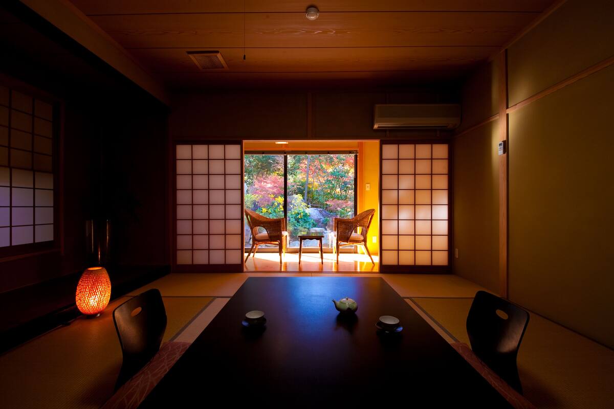 Habitació japonesa tradicional amb tatami, una taula baixa i joc de te, i portes corredisses que donen a un jardí de fulles colorades.