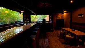 A dimly lit bar area with wooden furniture and a view of autumn foliage through large windows. - Kogane Sansou (Nakatsu)