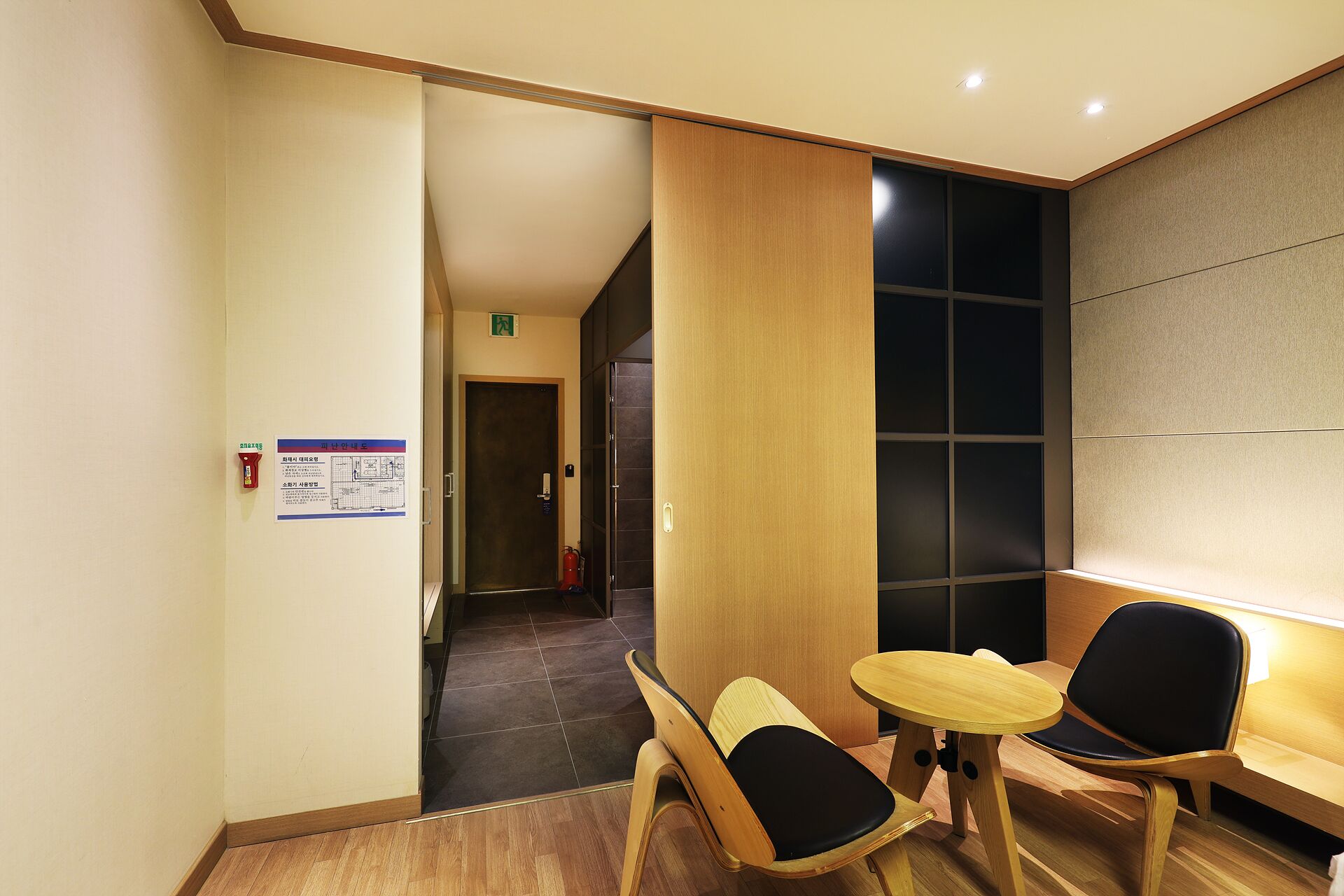 Un vestíbulo de hotel con un mostrador de recepción de madera, dos sillas de madera con cojines negros, una mesita redonda y un extintor colgado en la pared.