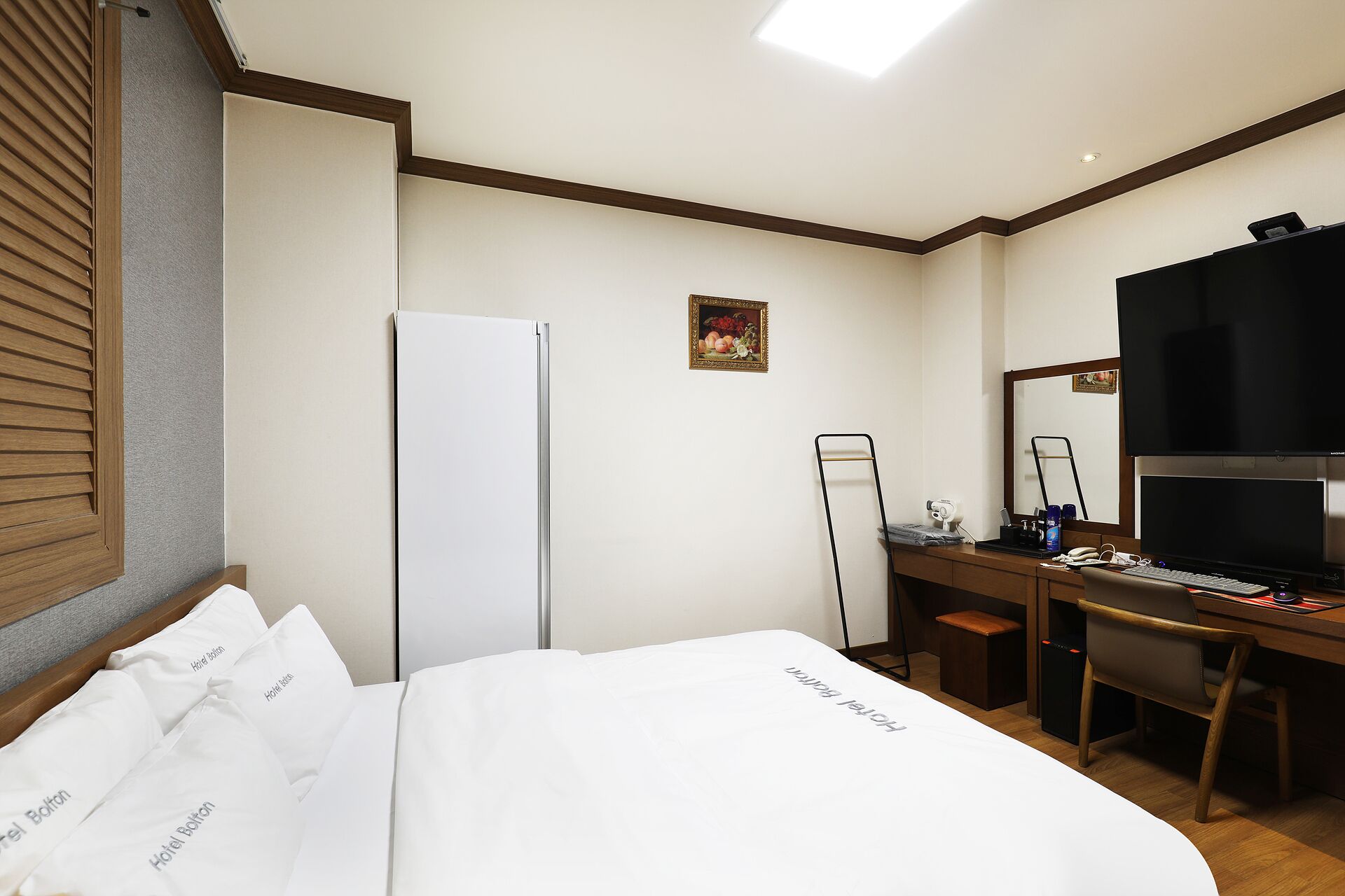 A hotel room with a bed, a desk, a refrigerator, a mirror, and a picture on the wall.