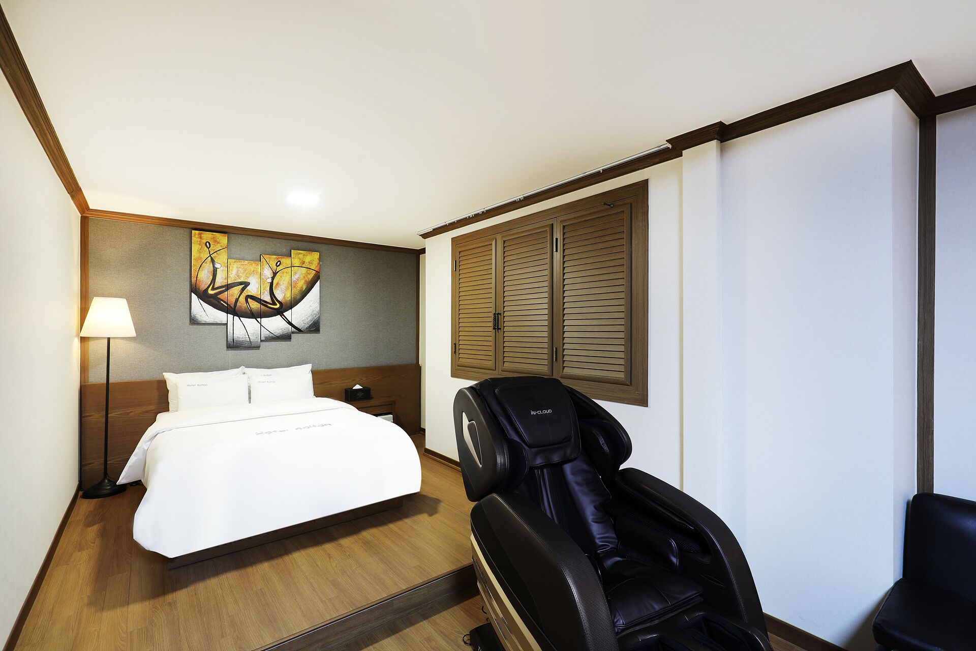 A hotel room with a bed, a massage chair, a painting, and a window with shutters.