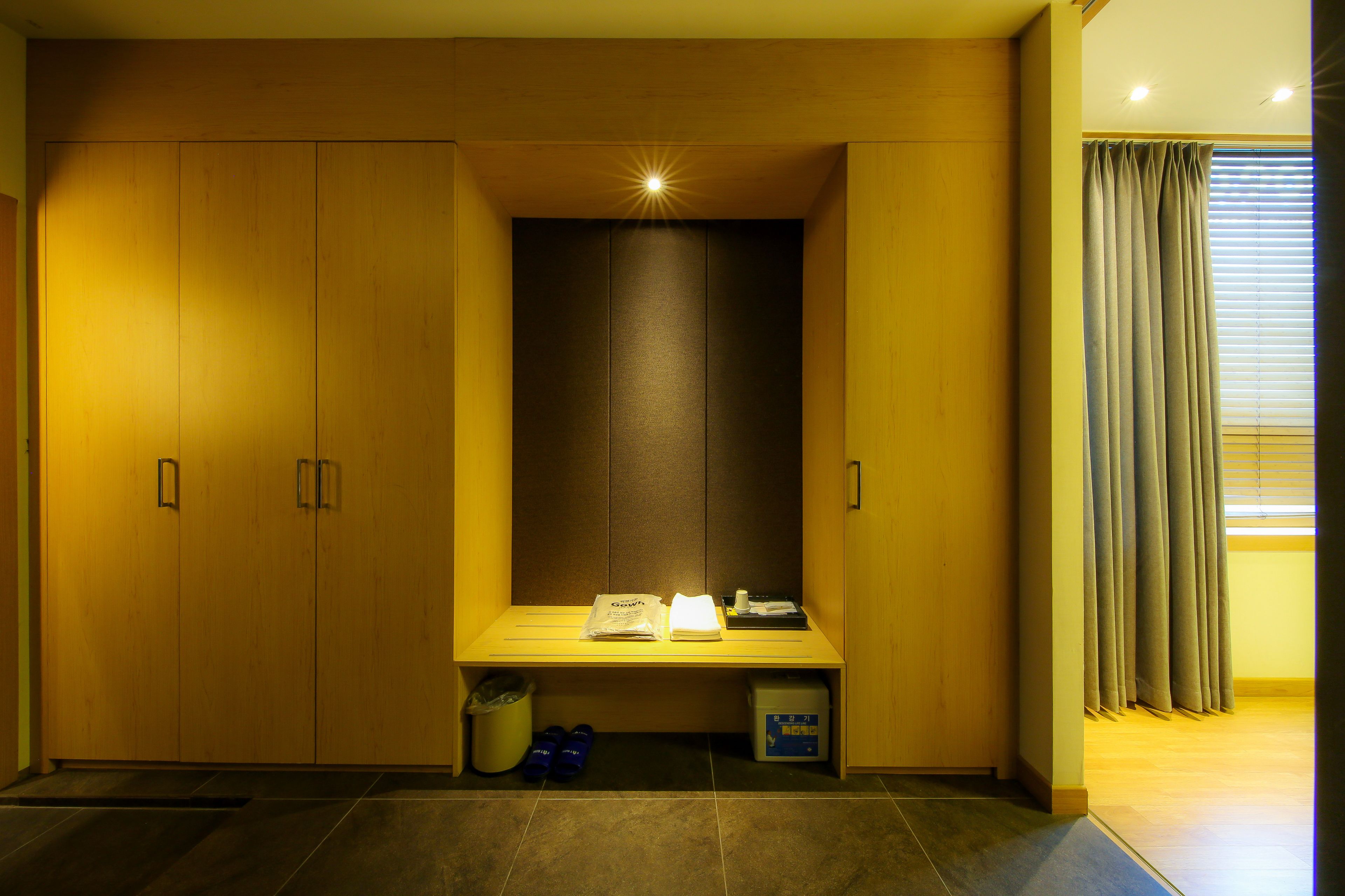 A hotel room with a wooden wardrobe, a bench with a small table, and a window with curtains.