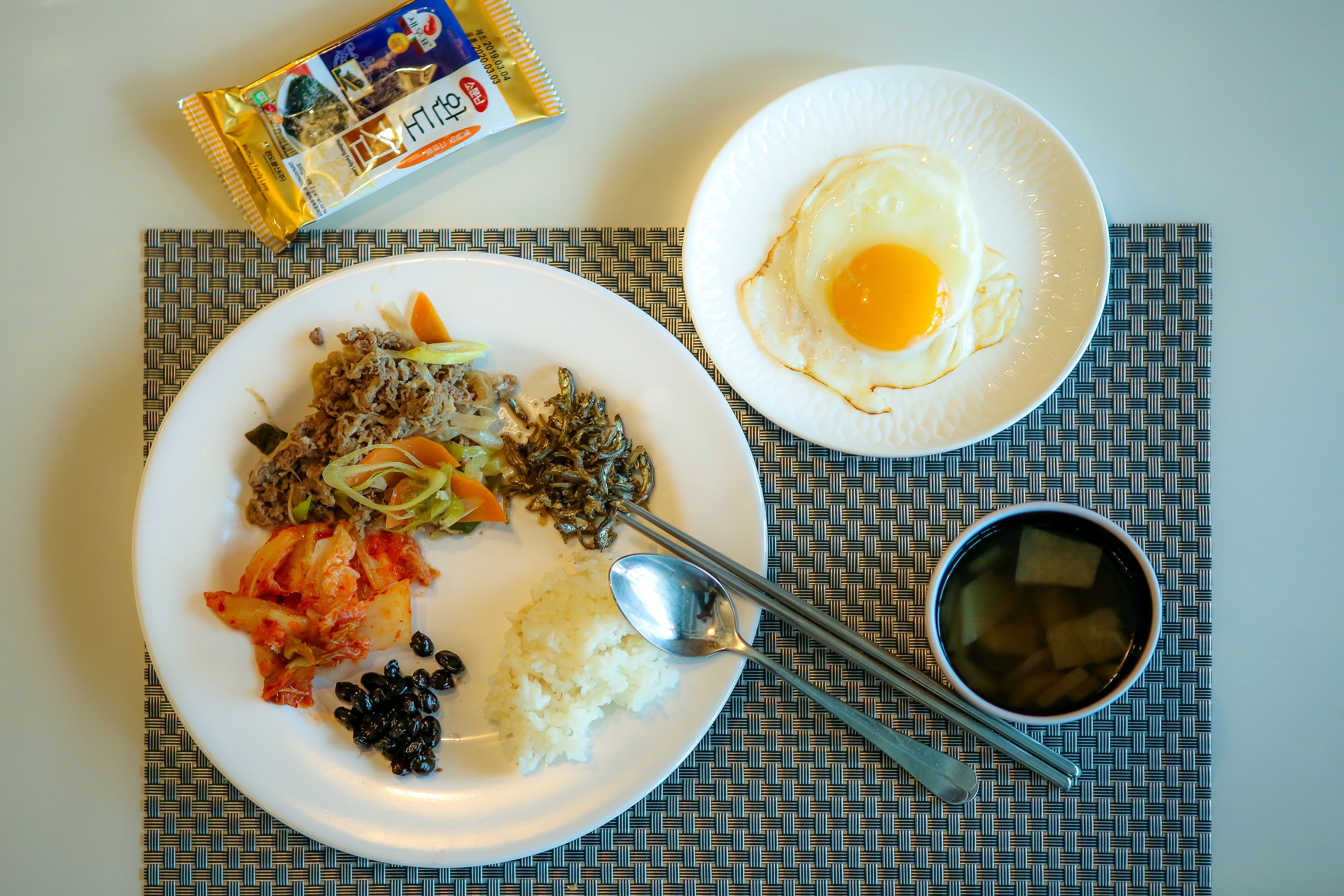 A meal with rice, kimchi, and a fried egg on a plate, accompanied by a packet of snacks and a bowl of soup.