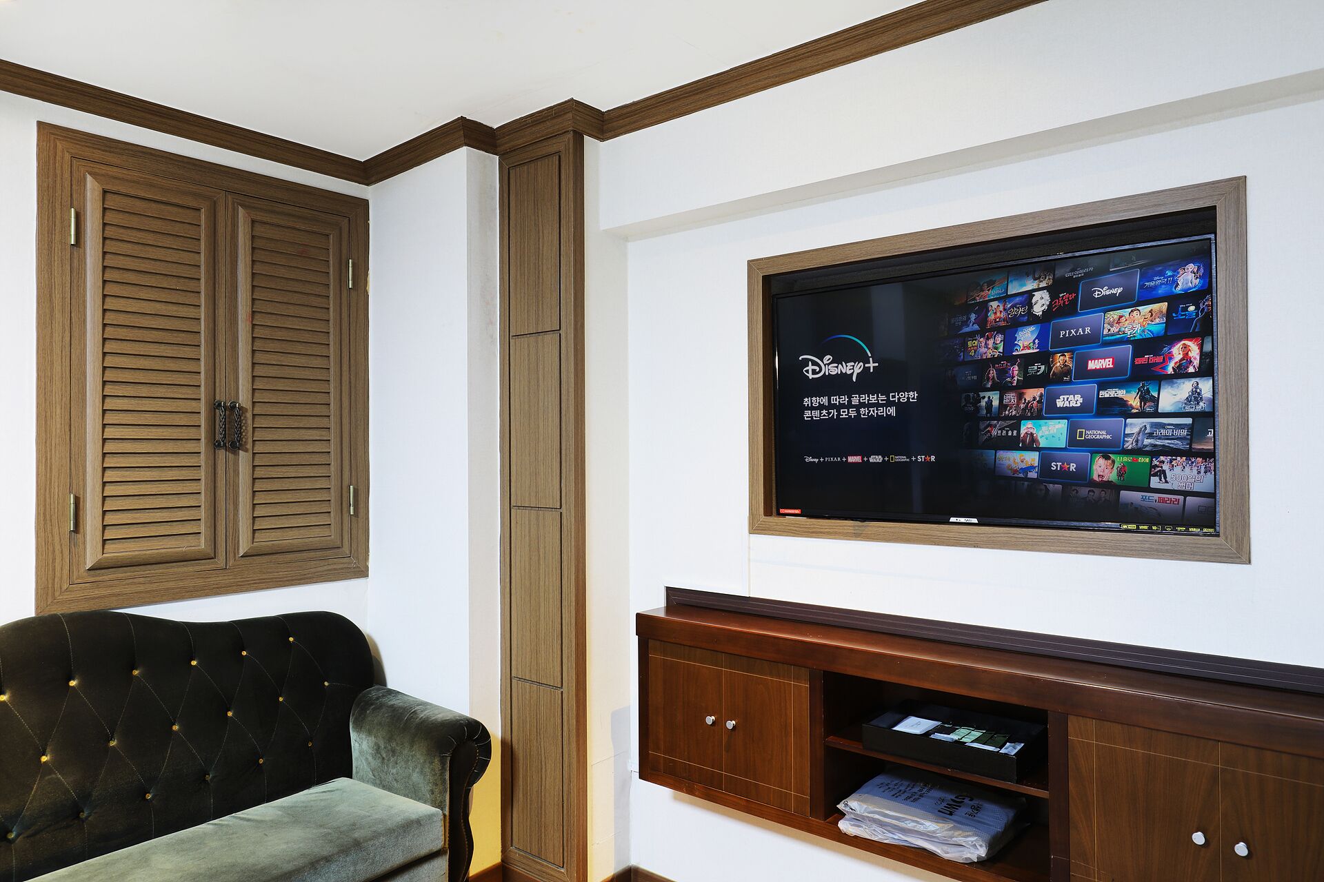 A living room with a television mounted on the wall displaying various streaming services. There is a wooden cabinet below the TV and a tufted armchair to the left.