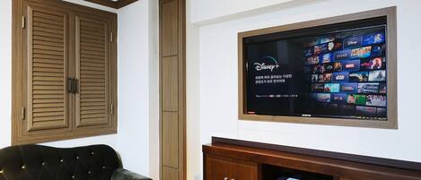 A living room with a television mounted on the wall displaying various streaming services. There is a wooden cabinet below the TV and a tufted armchair to the left.