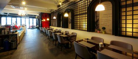 A restaurant with a long counter, multiple tables, and chairs. There are hanging lights and a wall with a grid pattern.