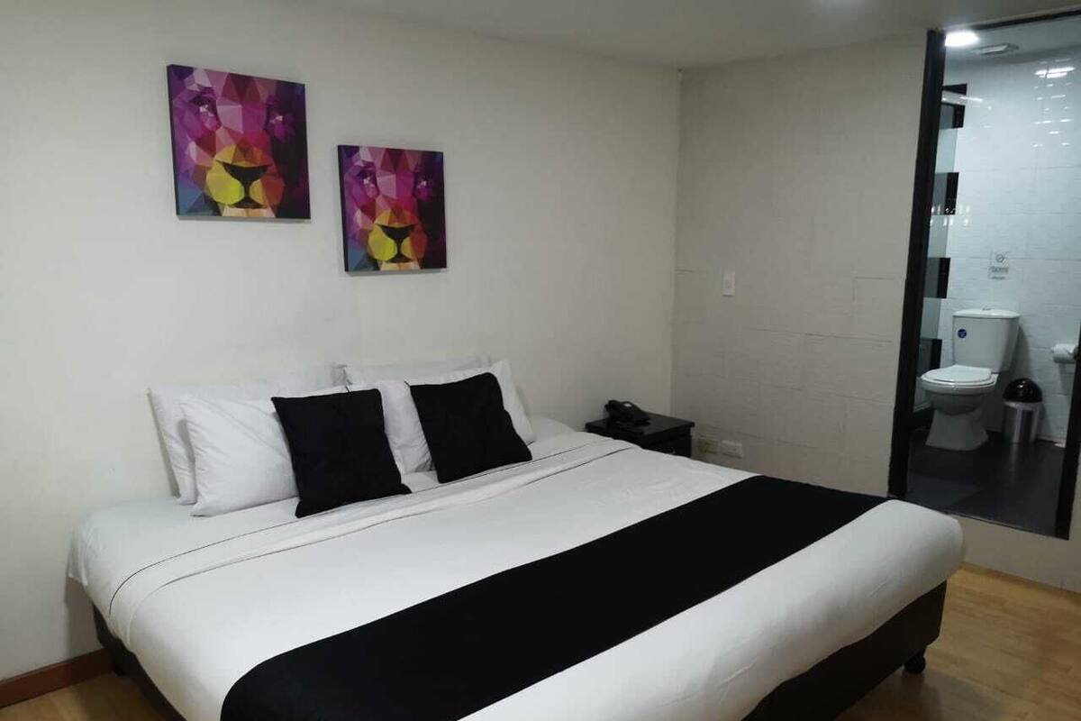 Una habitación de hotel con una cama grande, ropa de cama blanca y negra, dos cuadros abstractos en la pared y un baño visible con inodoro y lavamanos.