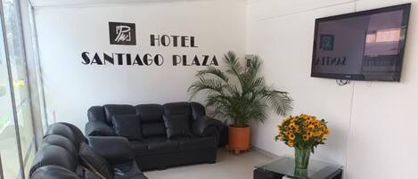 A hotel lobby with a black leather sofa, a potted plant, a vase with flowers, and a television mounted on the wall.