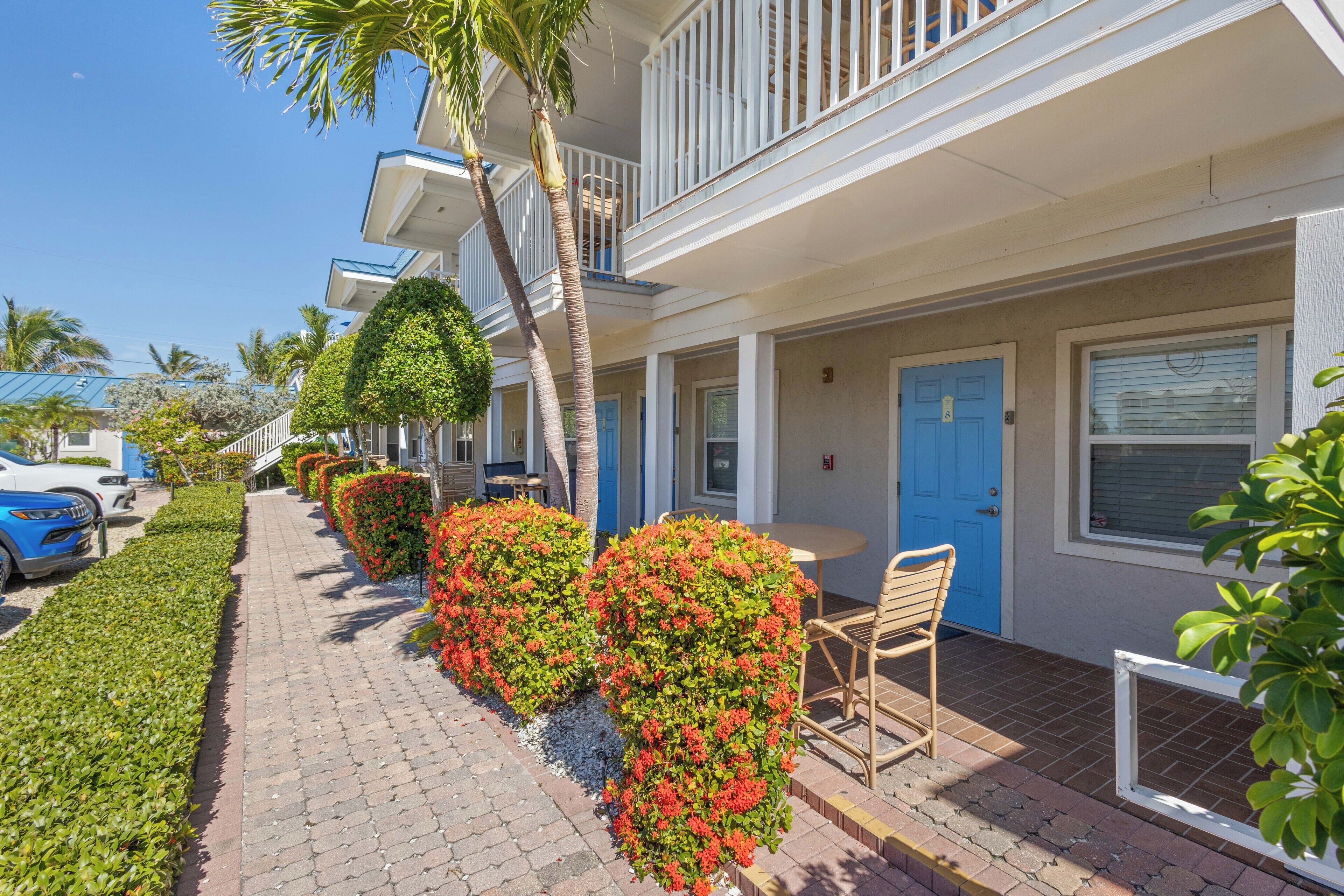 A row of hotel rooms with balconies, a paved walkway, and neatly trimmed bushes.