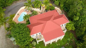 An aerial view of a large house with a red roof, surrounded by lush greenery and a swimming pool.
