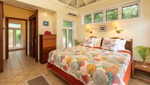 A bedroom with a large bed, a wooden dresser, a nightstand, and a window with three panes.