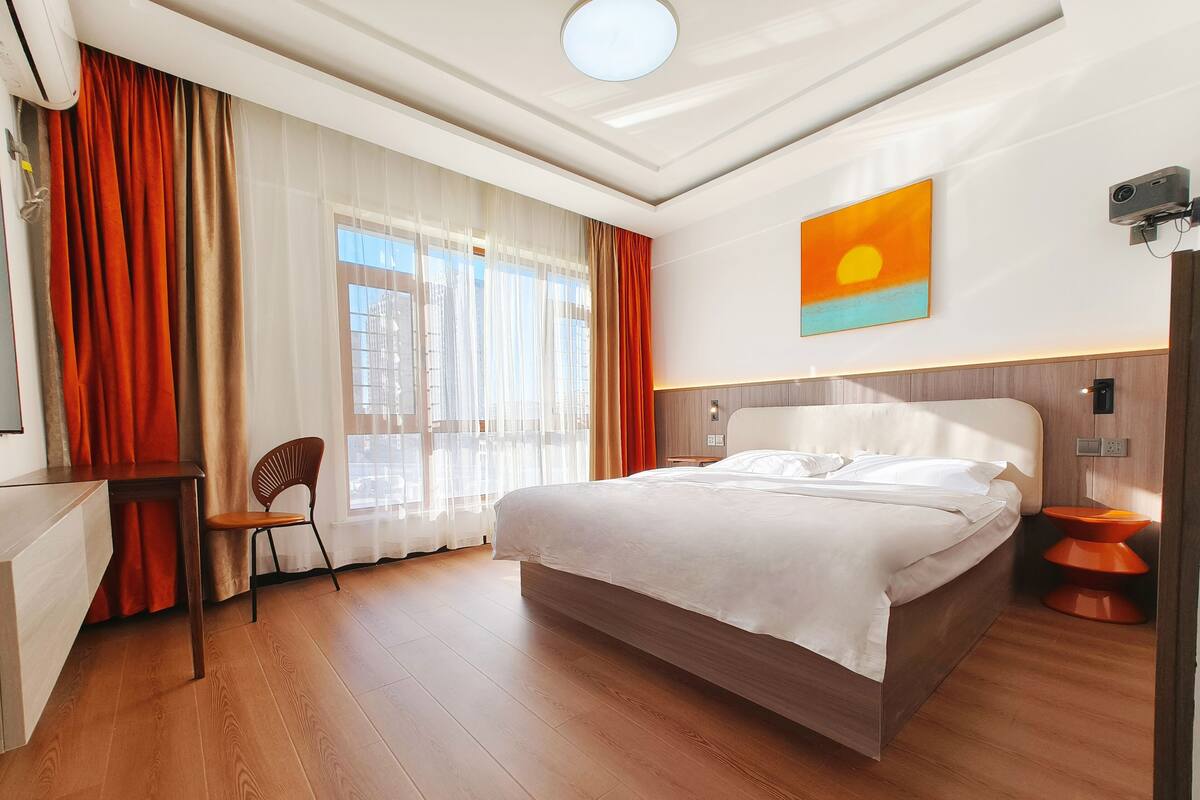 Ein Hotelzimmer mit einem großen Bett, einem Schreibtisch, einem Sessel und Blick auf die Stadt durch das Fenster.