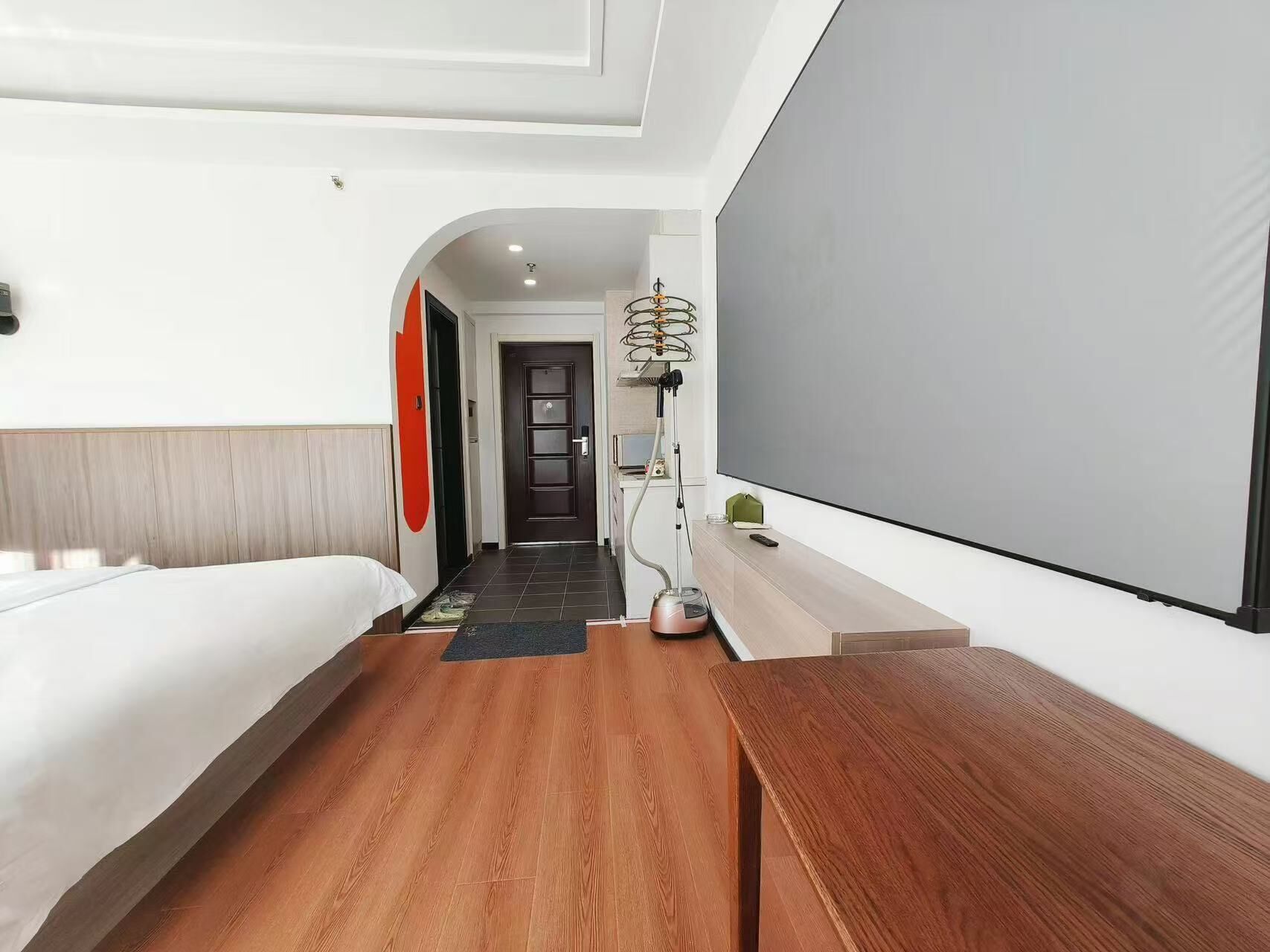 Habitación de hotel con cama, escritorio, televisión y un pasillo que lleva a una puerta.