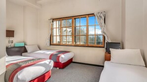 Habitación de hotel con dos camas, un escritorio con computadora, una ventana con cortinas y vistas a árboles y edificios.