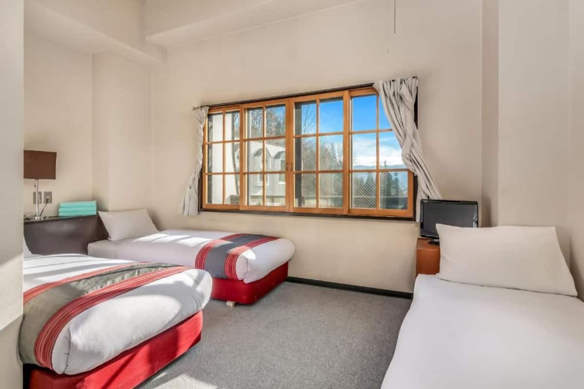 A hotel room with two beds, a desk with a computer, a window with curtains, and a view of trees and buildings outside.