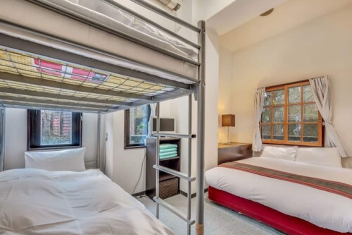 A hotel room with bunk beds, a window with curtains, a bedside table with a lamp, and a small shelf with folded towels.