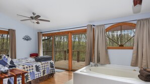 A bedroom with a large window, a ceiling fan, a bed with a checkered blanket, a wooden floor, and a bathtub.