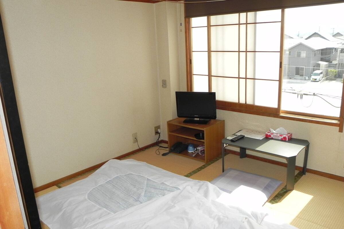 Une pièce avec un sol recouvert de tatamis, un téléviseur posé sur un meuble en bois, une petite table avec une boîte de mouchoirs, et une vue sur les maisons à travers la fenêtre.
